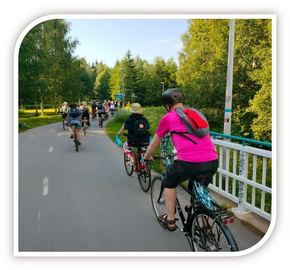 Useita pyöräilijöitä kesäisessä puistossa ylittämässä valkoista siltaa.